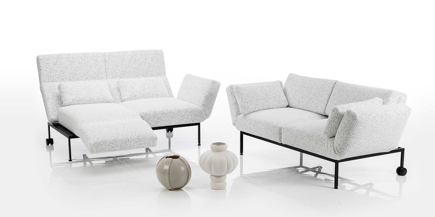 Kategorie roro medium Zwei Sofas mit weißen Stoffbezügen und schwarzem Untergestell. Die linke Armlehne des links stehenden Sofas ist zur Fußstütze vorgeschwenkt.