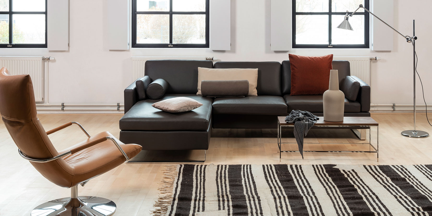 Sitz- und Decokissen von brühl Eine Eckcouch in schwarzem Leder. Auf der Couch liegen Kissen und Kissenrollen in verschiedenen Größen und in Stoff und Leder drapiert.