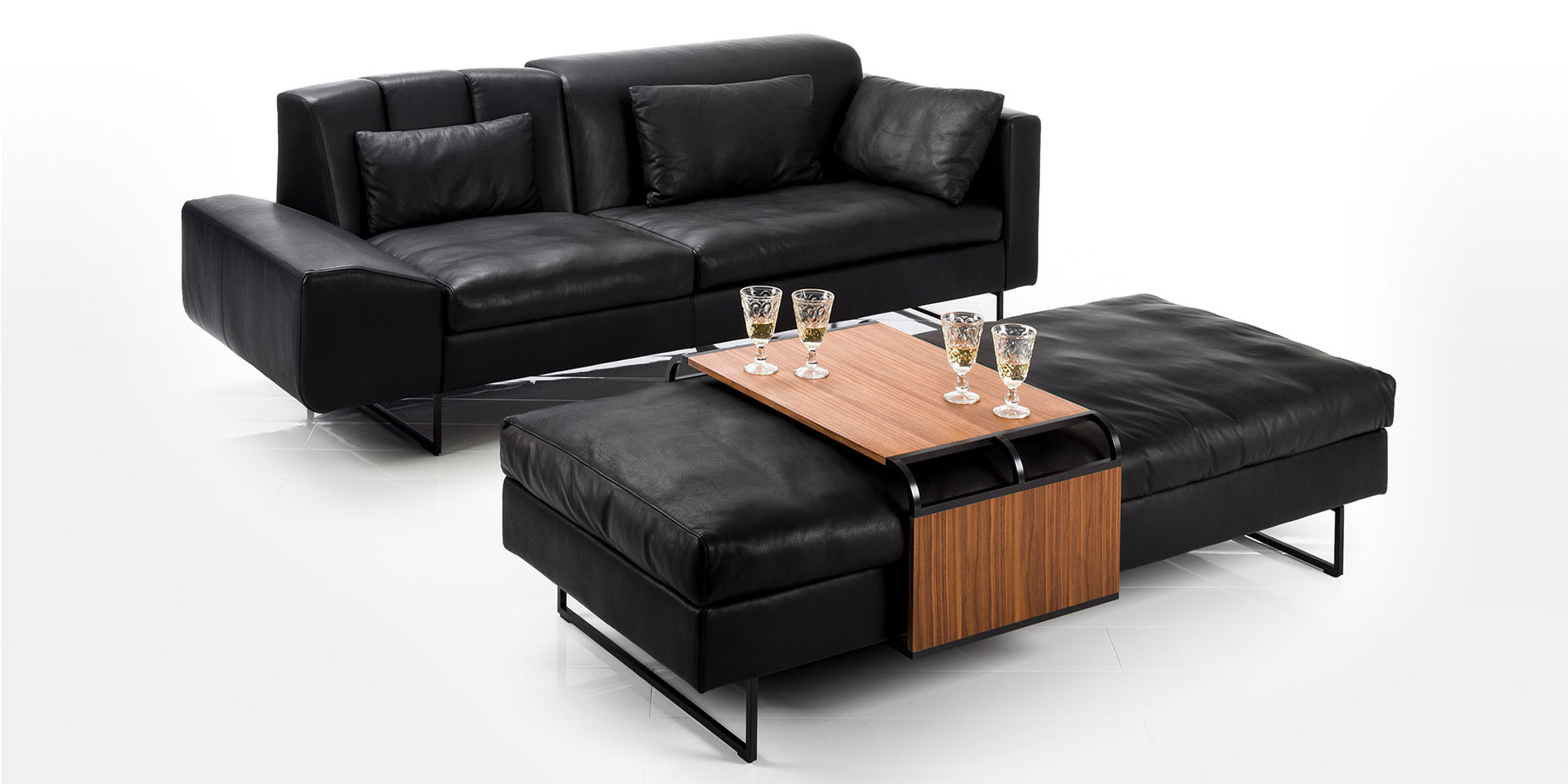 Bänke von brühl. Mit und ohne Rückenlehne. Eine schwarze Ledercouch und eine breite Sitzbank in schwarzem Leder. Auf der Bank liegt ein verschiebbarer Tisch aus dunklem Holz.