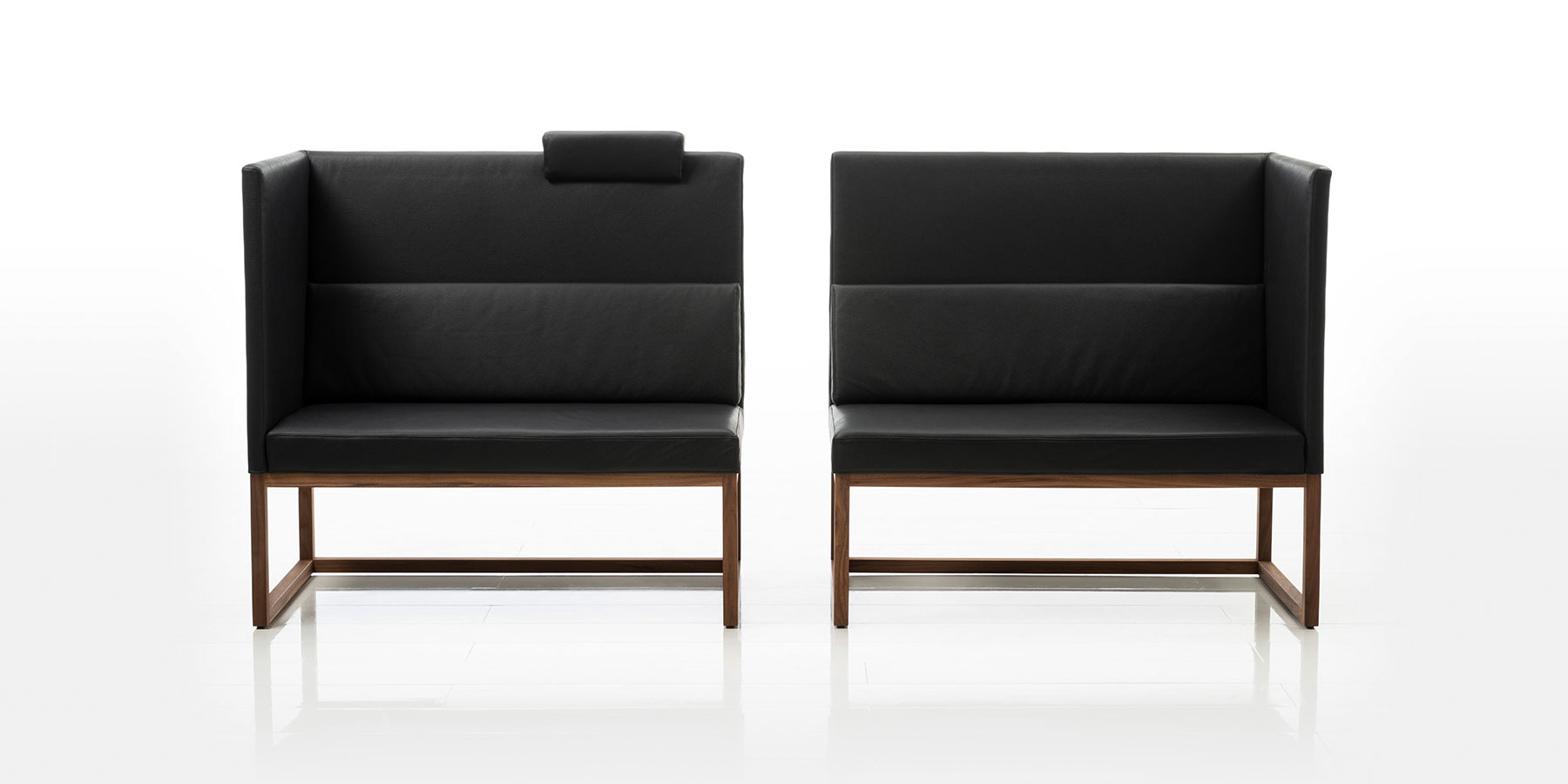 Zwei Sitzbänke in schwarzem Leder mit hoher Rücken- und Seitenlehne stehen mit geringem Abstand nebeneinander. Jede Sitzbank hat nur ein Seitenteil. Die einander gegenüberliegenden Seiten sind offen. Das Gestell ist aus dunklem Holz gefertigt.