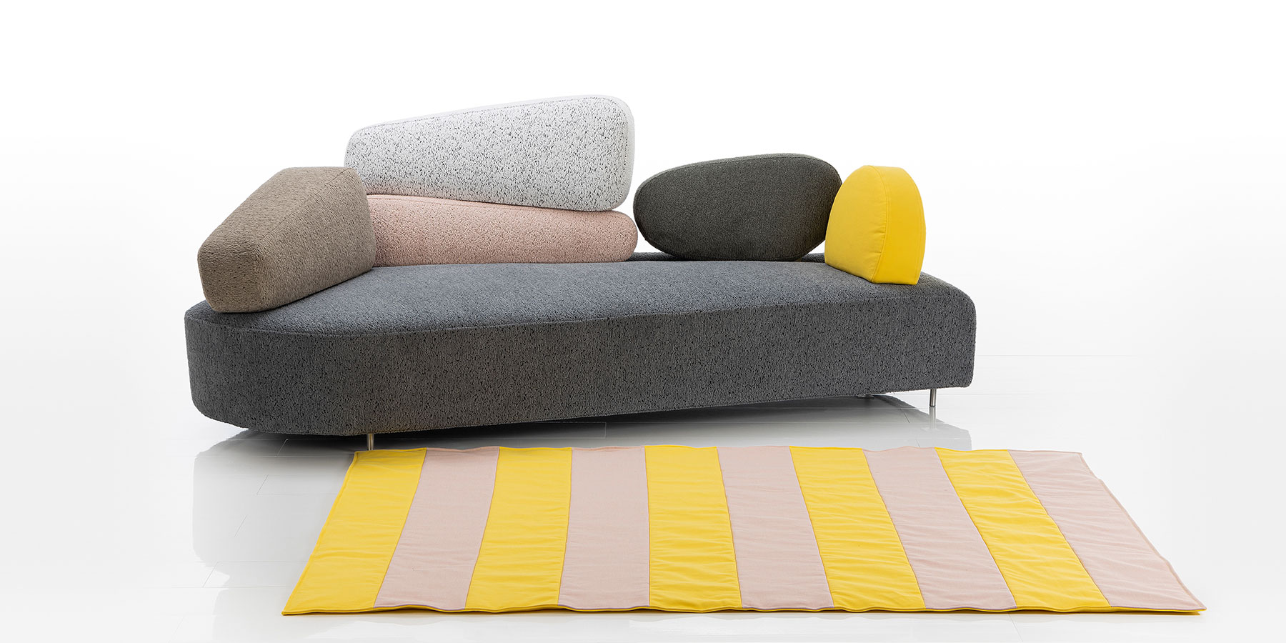 Sofa brühl mosspink Ein asymmetrisch geformtes Sofa mit einer Sitzbasis aus grauem Stoff. Rücken und Seiten bestehen aus sechs unregelmäßig geformten Segmenten in den Farben braun, rosé, hellgrau, dunkelgrau und gelb.