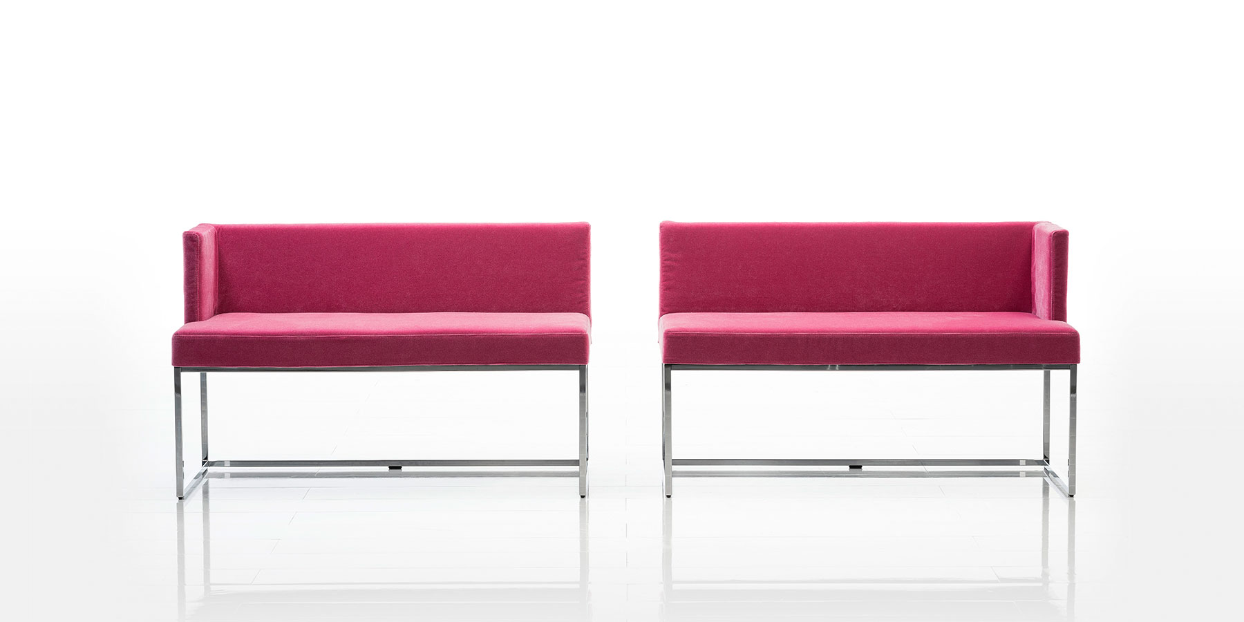 Zwei Sitzbänke mit niedriger Rückenlehne und in pinkem Stoff stehen mit geringem Abstand nebeneinander. Jede Sitzbank hat nur ein Seitenteil. Die einander gegenüberliegenden Seiten sind offen. Das Gestell ist aus Chrom glänzendem Edelstahl.