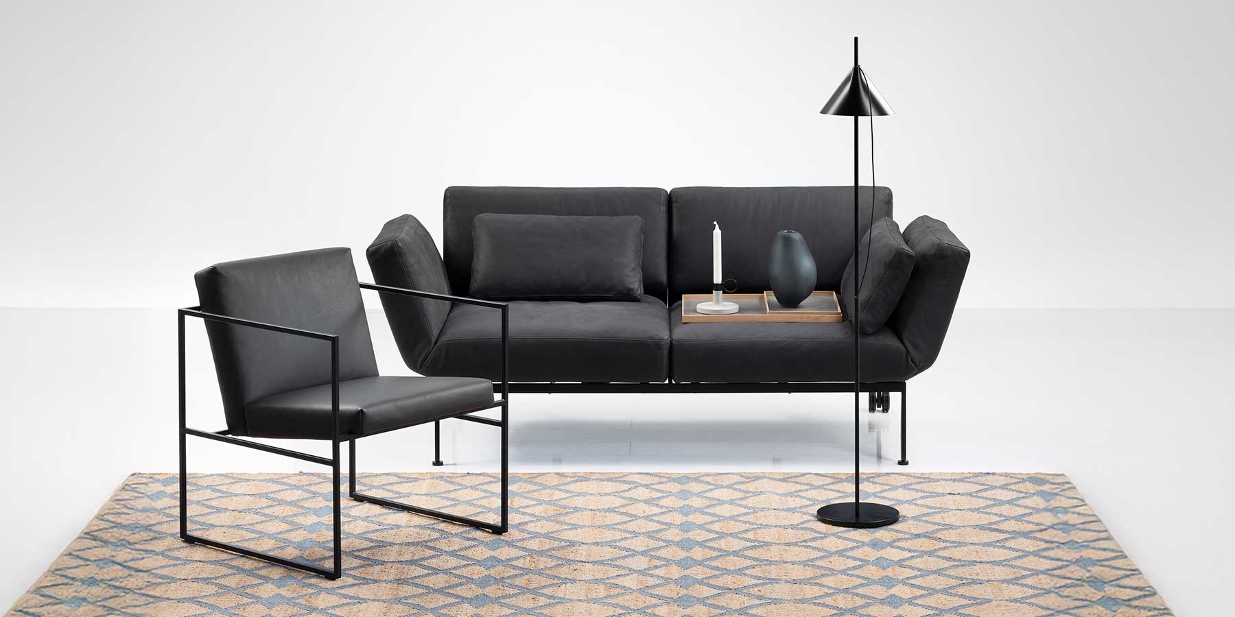 Multifunktionssofa roro medium soft. Schwarzes Ledersofa mit einem Rückenkissen. Auf der rechten Sitzfläche steht ein Tablett mit einer Kerze und einer Vase. Vor dem Sofa stehen eine schwarze grazile Stehlampe rechts und ein mit schwarzem Leder bezogener Stuhl.
