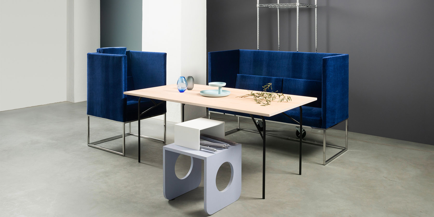 Ein Sessel und eine Sitzbank mit jeweils hohen Rückenlehnen und Seiten in einem sehr dunklem blau stehen um einen grazilen Esstisch mit einer Tischplatte in hellem Holz.
