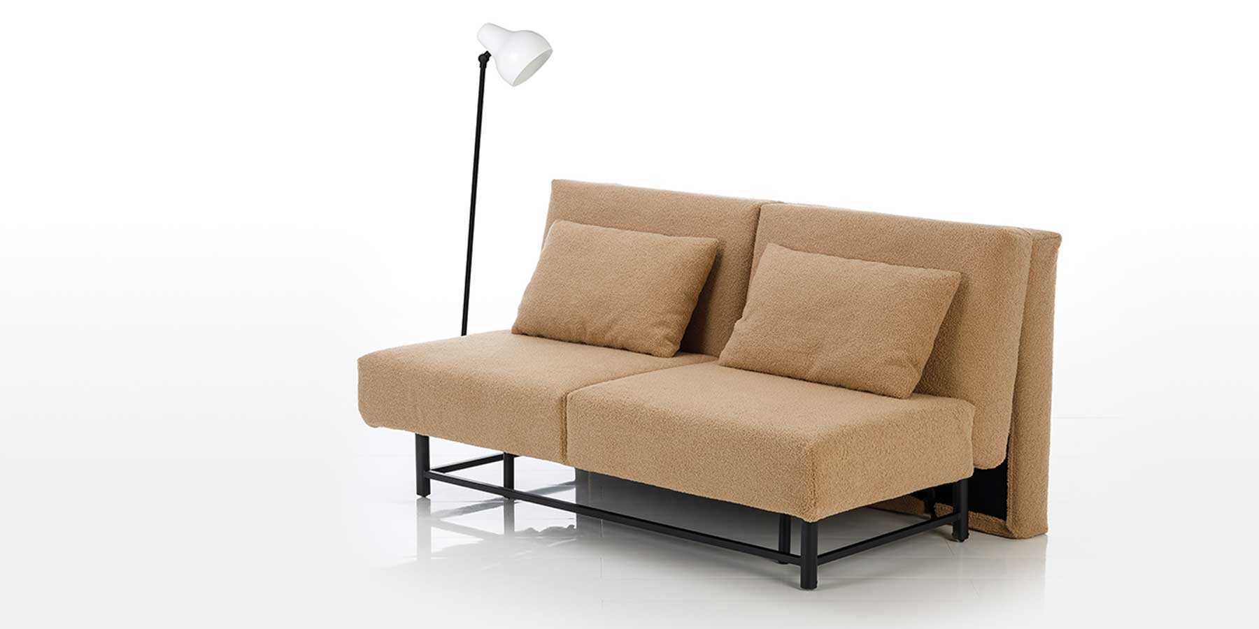 Bettsofa first class von brühl Beige-braunes Sofa mit zwei großen Rückenkissen und schwarzem Untergestell aus Metall. Links daneben steht eine Stehlampe.